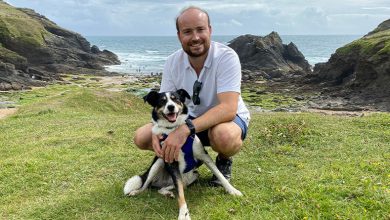 A man on a hillside near a beach cuddling a dog