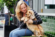 A woman cuddling a dog sat on a bench smiling.